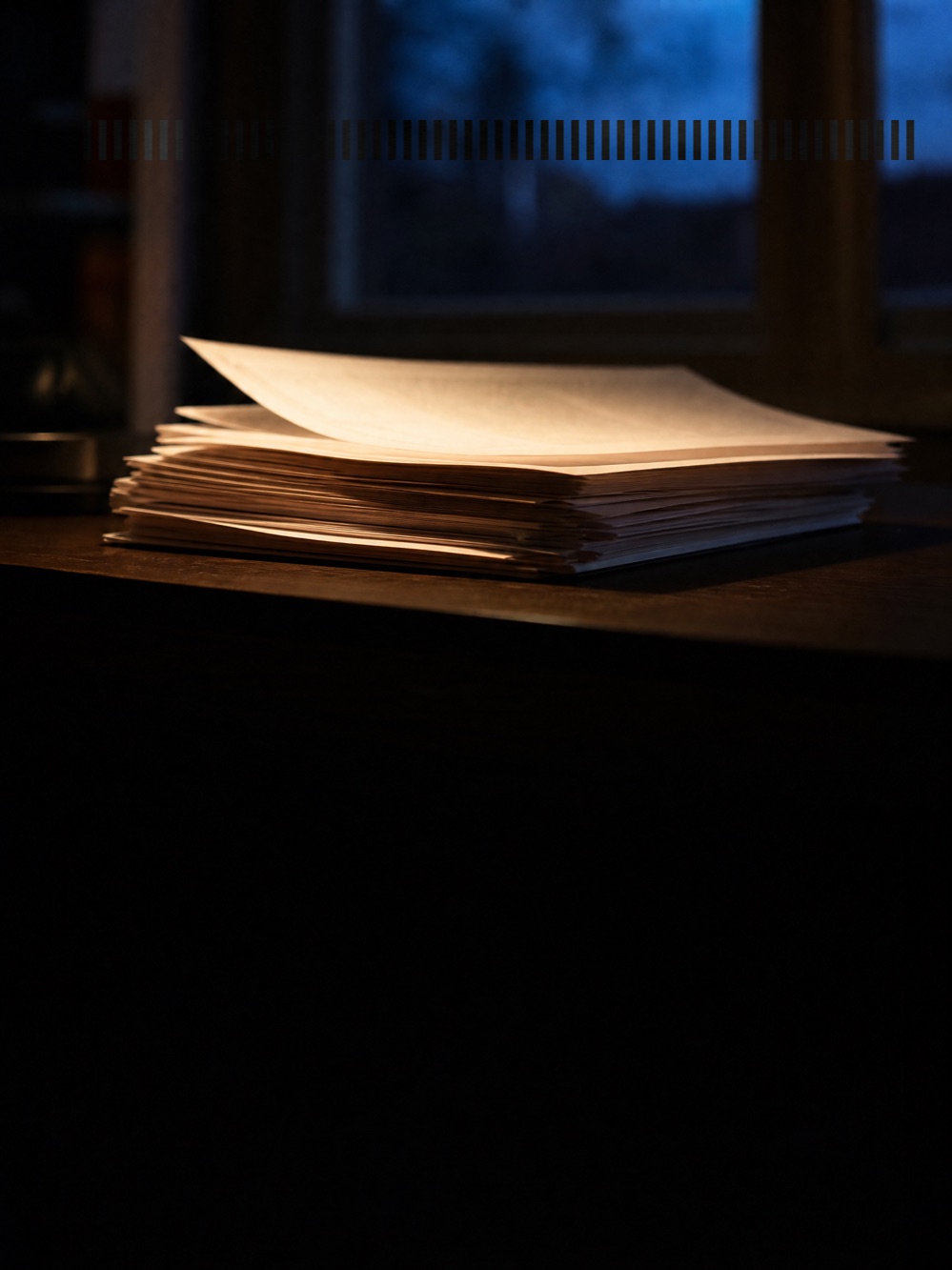 Ein Stapel loser Blätter auf einem dunklen Schreibtisch, das oberste Blatt leicht angehoben, sodass die Lagen darunter der Reihe nach sichtbar werden; warmes Lampenlicht streift die Kanten, dahinter ein Fenster in der blauen Stunde.